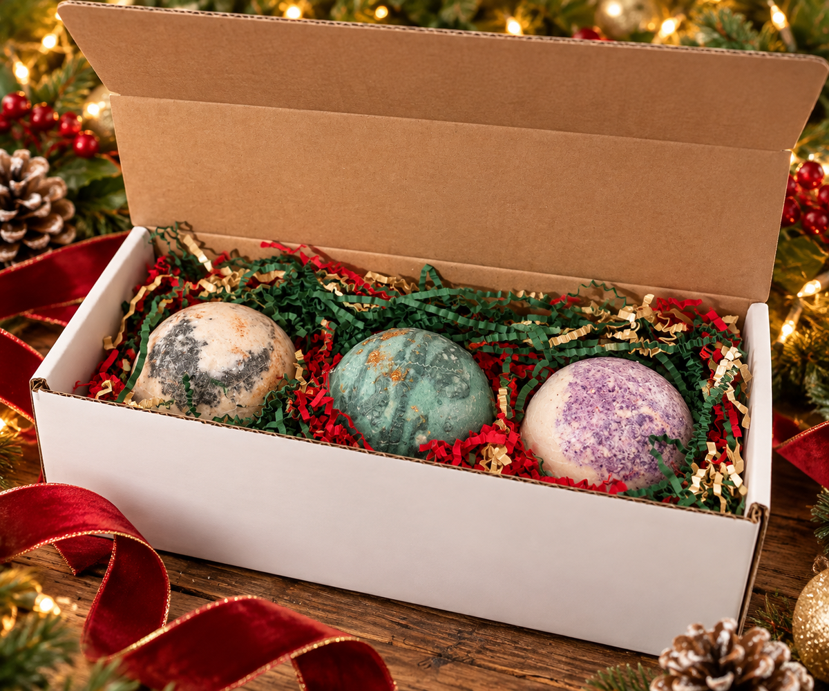 Three artisan bath bombs nestled in festive holiday fill in a gift box, surrounded by greenery and warm holiday lights
